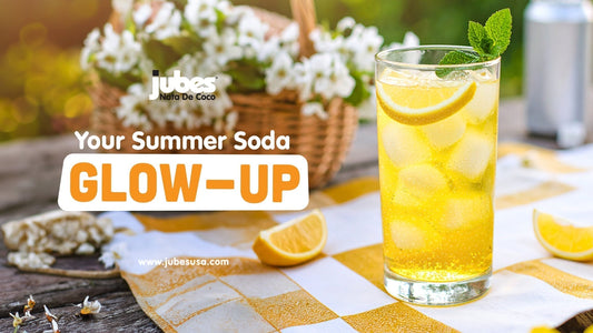 Glass of golden soda with ice cubes, lemon slices, and a mint garnish placed on a checkered cloth outdoors, with text that reads 'Your Summer Soda Glow-Up'.