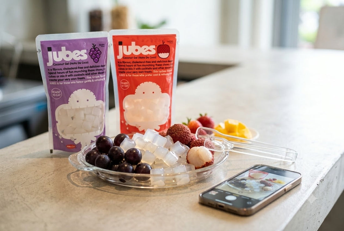Purple Grape and red Lychee Jubes Nata De Coco pouches on a kitchen counter beside a bowl of grapes, lychee, and coconut jelly cubes.