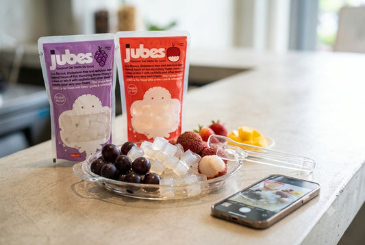 Purple Grape and red Lychee Jubes Nata De Coco pouches on a kitchen counter beside a bowl of grapes, lychee, and coconut jelly cubes.