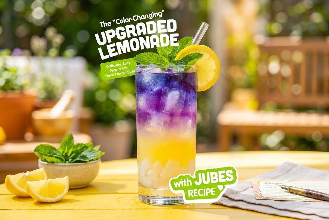 Tall glass of purple-to-gold color-changing lemonade with ice, mint, and a lemon wheel on a sunny outdoor table, Jubes nata de coco settled at the bottom.