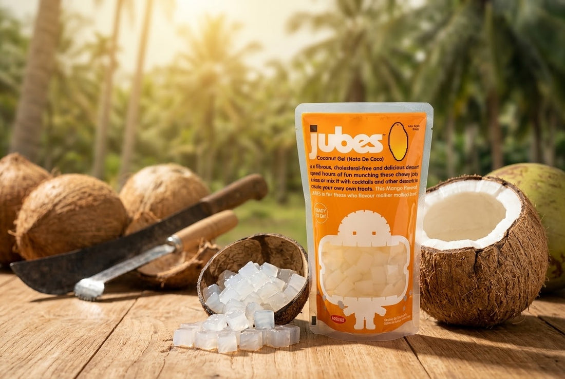 A Jubes Mango coconut jelly pouch on a wooden table in a coconut grove, next to a split coconut, a whole coconut, a machete, and a bowl of coconut cubes.