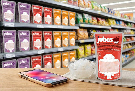 Jubes Lychee coconut jelly pouch and a bowl of coconut gel cubes on a counter, with a smartphone, backed by a shelf stocked with Jubes flavors.