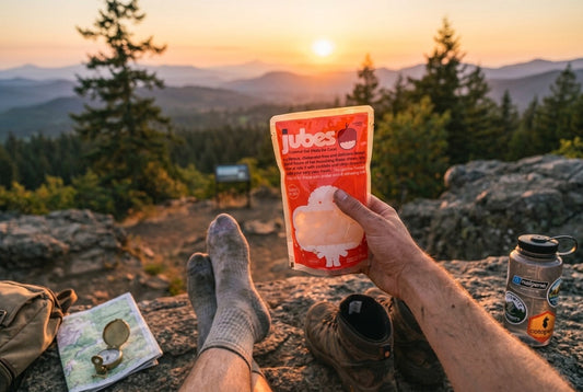 Hiker's feet in wool socks rest on a rock at sunset, holding a Jubes coconut jelly pouch, with hiking boots, a compass, and a water bottle nearby.