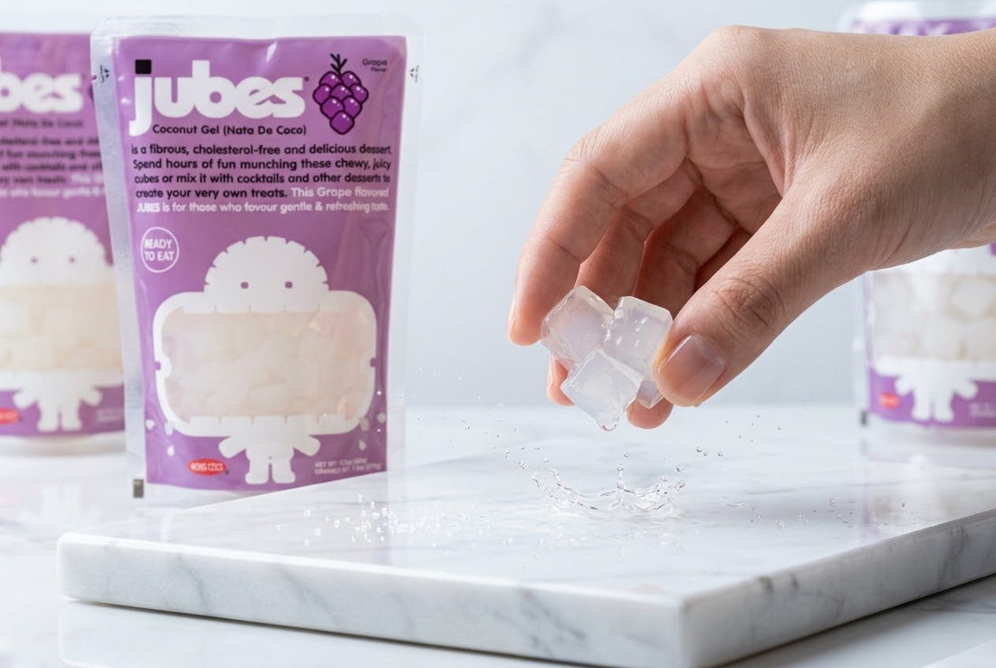 A hand drops bouncy Jubes coconut jelly cubes into a splash of water, with grape-flavored Jubes pouches softly blurred in the background.