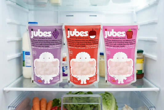 Three Jubes coconut jelly pouches, grape, lychee, and strawberry, standing on a fridge shelf next to milk, yogurt, and condiments.