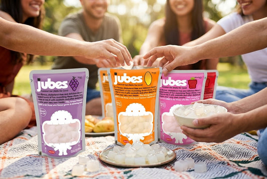 Friends at an outdoor picnic reaching for Jubes coconut jelly pouches in grape, mango, and strawberry, with a bowl of loose jelly cubes.