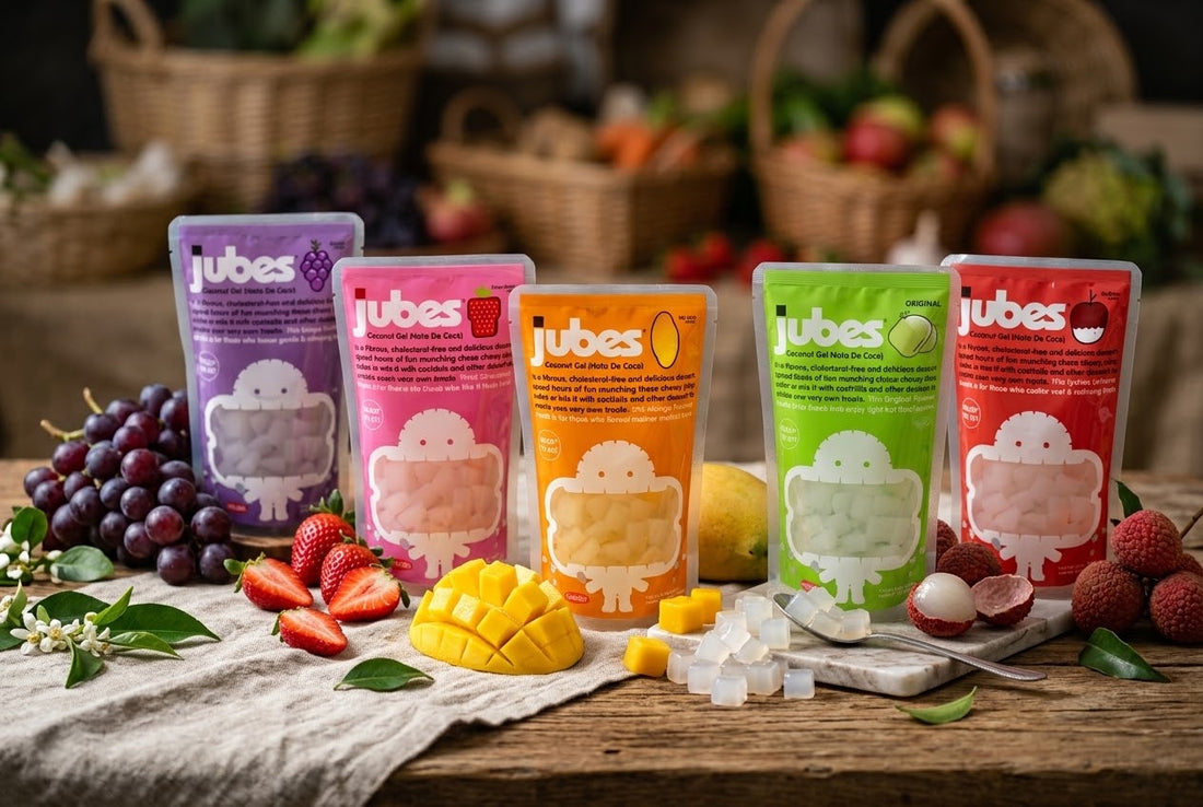 Five Jubes coconut jelly pouches in grape, strawberry, mango, original, and lychee lined up with fresh fruit on a rustic wood table.