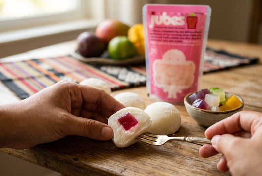A halved mochi ball revealing a pink jelly cube filling, held over more mochi balls, a bowl of coconut jelly cubes, and a Jubes pouch.