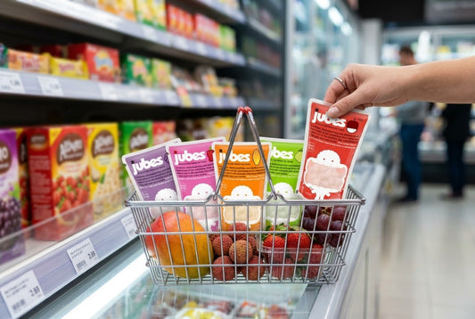 A hand grabs a red Jubes pouch to add to a grocery basket already holding four Jubes flavors and fresh fruit in a store aisle.