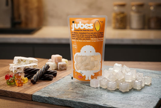 A Jubes Mango coconut jelly pouch on a counter next to nougat, gummy bears, licorice twists, and a pile of loose coconut jelly cubes.