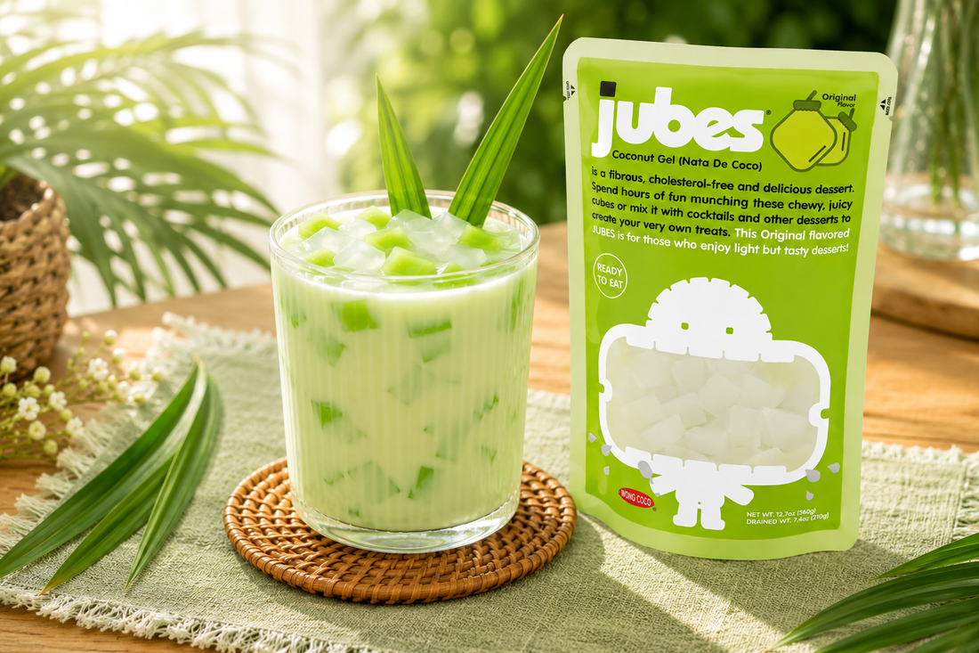 A green pouch of Jubes Coconut Gel next to a glass of creamy green drink with jelly cubes and pandan leaves, set on a sunlit table.
