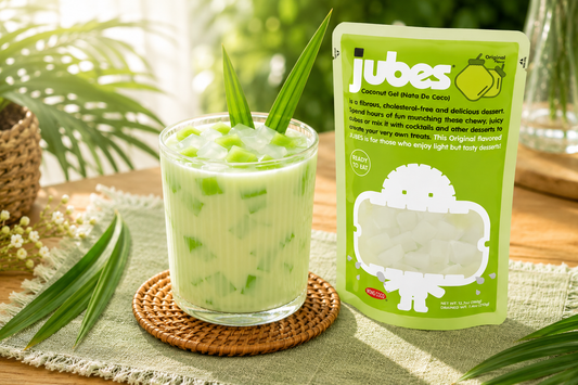 A green pouch of Jubes Coconut Gel next to a glass of creamy green drink with jelly cubes and pandan leaves, set on a sunlit table.