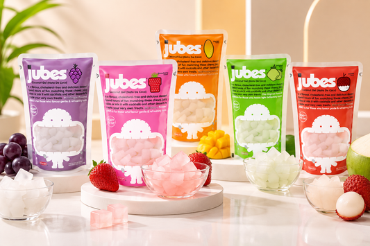 Five Jubes coconut jelly pouches in grape, strawberry, mango, original, and lychee with cube bowls and fresh fruit.