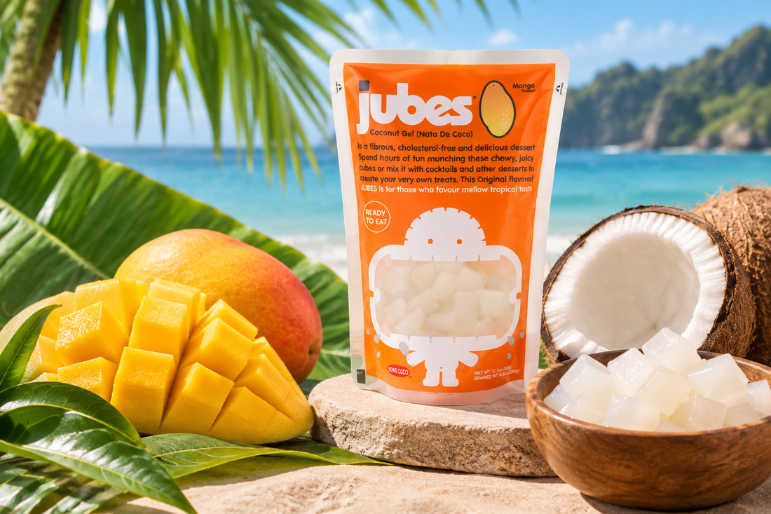 An orange Jubes Mango pouch on a beach rock beside fresh mango, a split coconut, and a bowl of coconut jelly cubes.