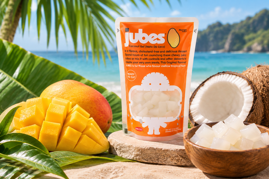 An orange Jubes Mango pouch on a beach rock beside fresh mango, a split coconut, and a bowl of coconut jelly cubes.