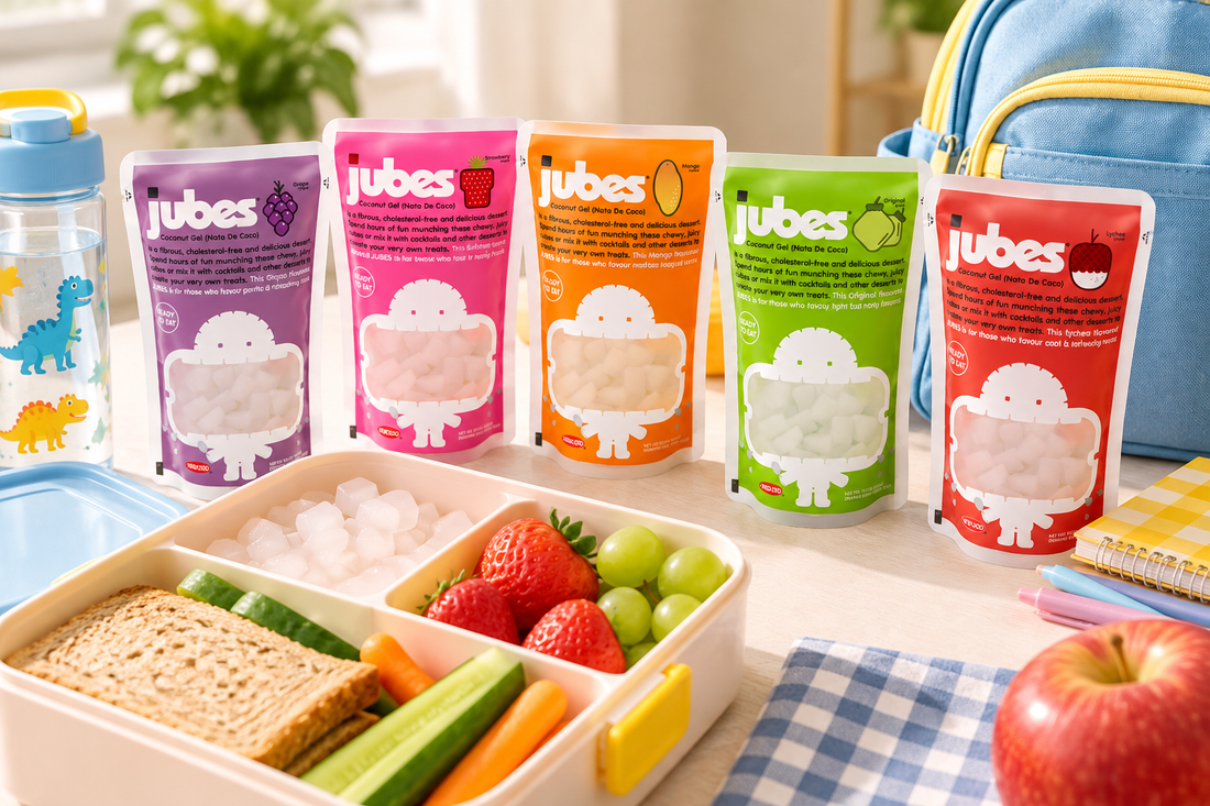 Five Jubes coconut jelly pouches in grape, strawberry, mango, original, and lychee beside a kids school lunchbox with fruit and veggies.