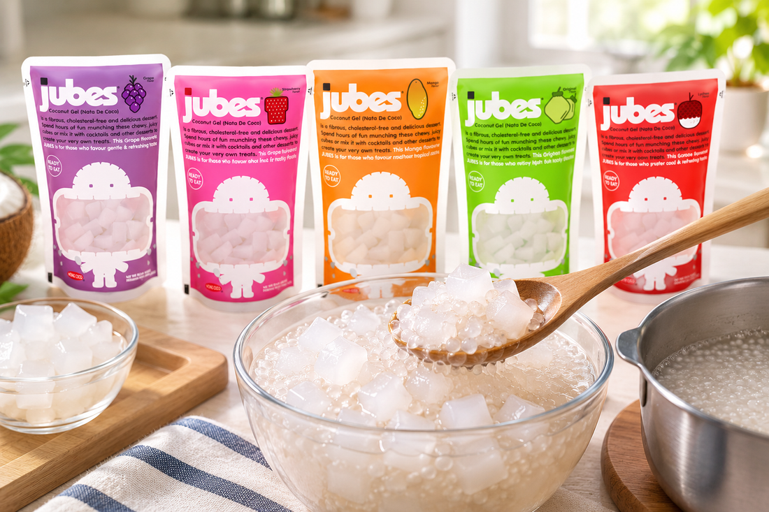 Five Jubes coconut gel pouches behind a glass bowl of cooked sago pearls and nata de coco cubes, with a wooden spoon and a pot of pearls.