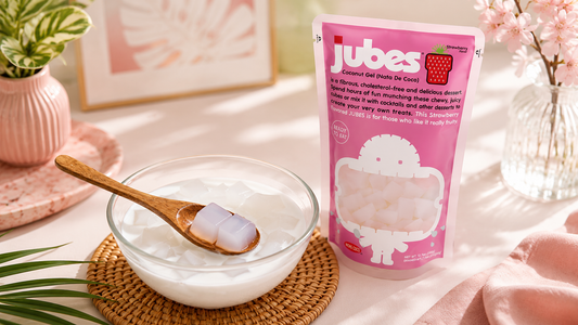A pink stand-up pouch of Jubes Strawberry Flavor Coconut Gel (Nata De Coco) next to a glass bowl filled with milky dessert and translucent gel cubes, with a wooden spoon holding two cubes.