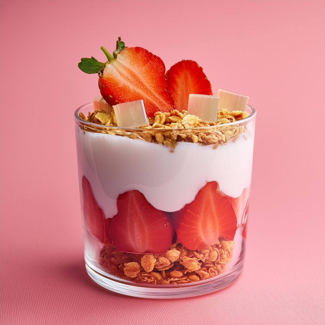 Glass of yogurt parfait layered with strawberries, granola, and coconut jelly cubes against a pink background.