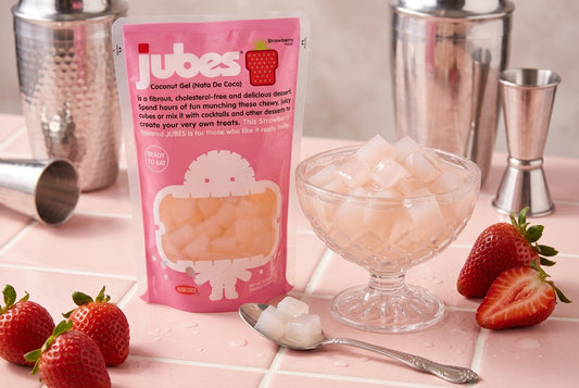 A pink stand-up pouch of Jubes Strawberry Flavor Coconut Gel (Nata De Coco) next to a clear glass dessert bowl filled with translucent, square coconut gel cubes. Fresh strawberries are scattered on a pink tiled countertop.