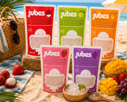 Jubes nata de coco coconut jelly pouches arranged on a sunny beach towel with tropical fruits, seashells, and ocean waves in the background, showcasing a refreshing summer snack.