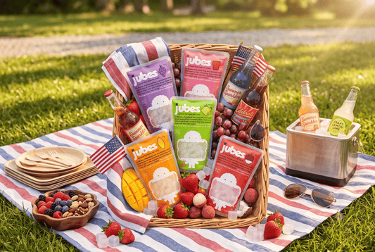 A 4th of July picnic setup on the grass featuring a wicker basket filled with various flavors of Jubes Nata De Coco pouches, sodas, fresh fruits, and small American flags on a red and blue striped blanket.