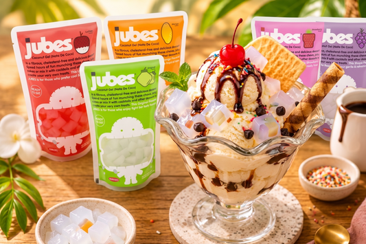 Vanilla ice cream sundae topped with chocolate syrup, cherries, and chewy nata de coco jelly cubes beside a pink Jubes coconut gel pouch on a tropical wooden table.