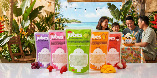A row of colorful Jubes Nata de Coco pouches on a white table, surrounded by fruits like grapes and strawberries. In the background, people are laughing at a beachside patio.