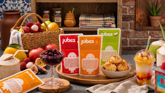 beach dessert spread featuring bowls of tropical fruit, shaved ice, coconut desserts, and Jubes nata de coco jelly cubes on a beach towel beside the ocean.