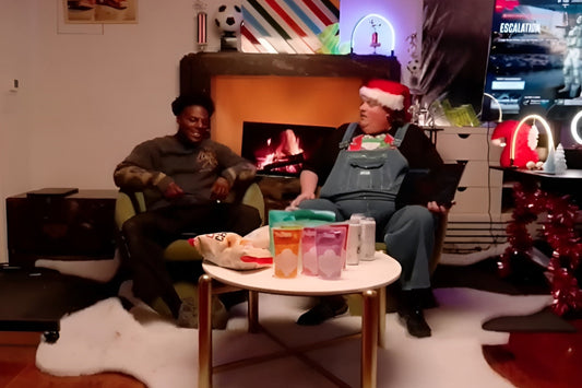Two popular live streamers, IShowSpeed and Clix dressed in a Santa hat and overalls, sitting together in a cozy room decorated for the holidays while holding a live stream session with snacks and drinks on the table.