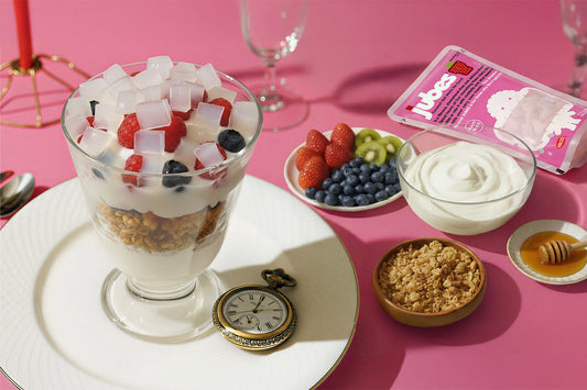 A yogurt parfait topped with berries and Jubes nata de coco, surrounded by fruit, granola, honey, and a Jubes pouch on a pink background.