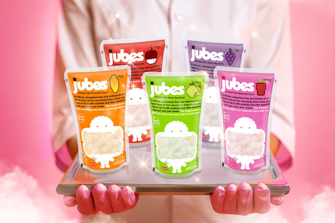 Person in a white coat holding a tray with five colorful Jubes nata de coco pouches in mango, lychee, grape, apple, and strawberry flavors against a pink background.