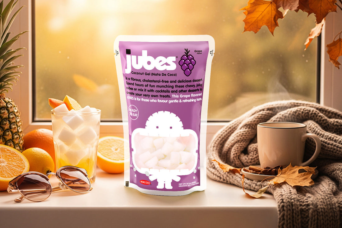 Purple Jubes package displayed on a windowsill with a glass of coconut jelly, surrounded by oranges, pineapple, a coffee cup, and autumn leaves in the background.