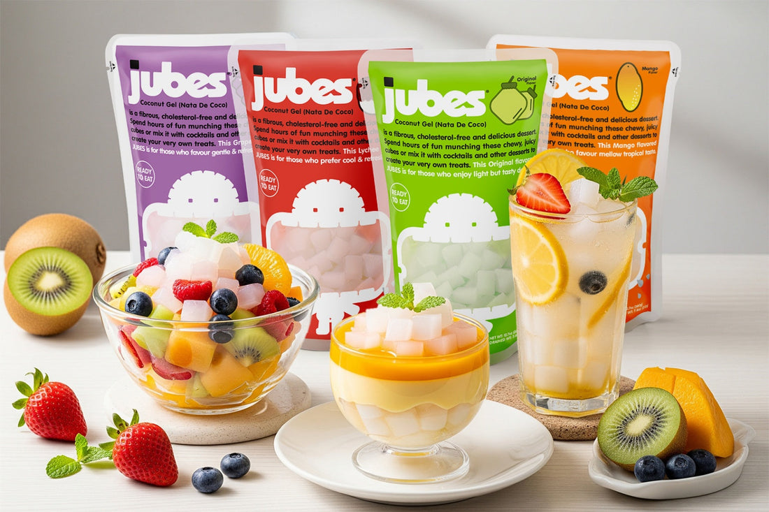Four Jubes nata de coco packs in different flavors displayed behind a fruit salad, parfait, and drink topped with coconut jelly cubes, with fresh fruit like strawberries, blueberries, and kiwi around them.