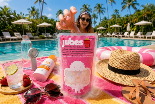 A smiling woman by a swimming pool reaches for a pink pouch of Jubes Strawberry Flavor Coconut Gel resting on a colorful towel alongside summer essentials like sunscreen, sunglasses, and a straw hat.