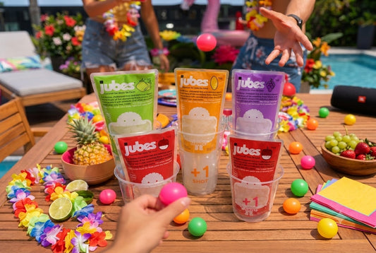 People playing a party game by tossing colorful plastic balls into clear cups holding various flavors of Jubes Nata De Coco pouches on a wooden poolside table.