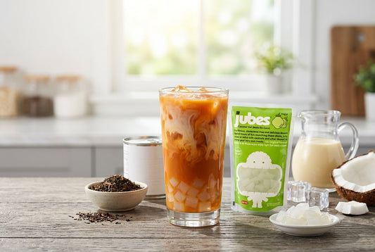 A tall glass of iced Thai tea with milk and translucent coconut gel cubes at the bottom, placed on a wooden kitchen counter next to a green pouch of Jubes Original Nata De Coco.
