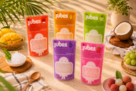 Five colorful stand-up pouches of Jubes Nata De Coco in various fruit flavors arranged together, surrounded by fresh fruits like mango, coconut, grapes, and lychee on a bright, sunny surface.