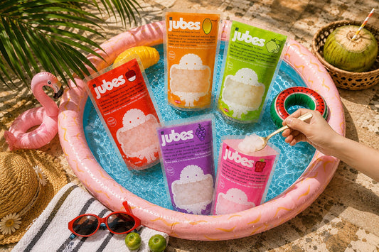 Five colorful pouches of Jubes Nata De Coco in various fruit flavors arranged inside a small, water-filled pink inflatable pool, creating a vibrant, summer-themed display.