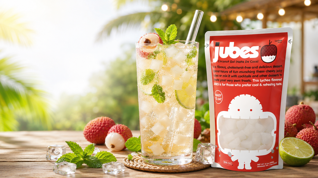 A red pouch of Jubes Lychee Flavor Coconut Gel next to a tall, refreshing iced drink filled with translucent nata de coco cubes, lime slices, mint leaves, and a glass straw on a sunny wooden table.