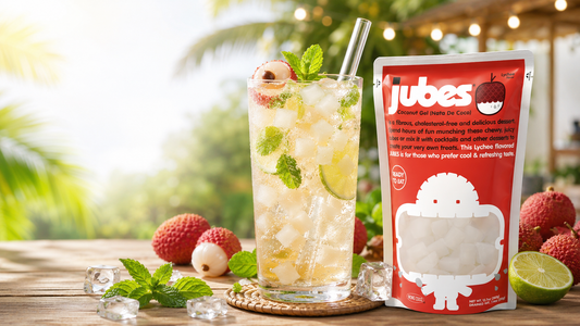 A red pouch of Jubes Lychee Flavor Coconut Gel next to a tall, refreshing iced drink filled with translucent nata de coco cubes, lime slices, mint leaves, and a glass straw on a sunny wooden table.