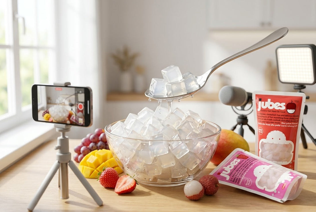 A content creation setup filming translucent coconut gel cubes being scooped from a glass bowl with a spoon, surrounded by fresh fruits and Jubes Nata De Coco pouches.