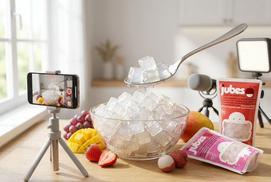A content creation setup filming translucent coconut gel cubes being scooped from a glass bowl with a spoon, surrounded by fresh fruits and Jubes Nata De Coco pouches.