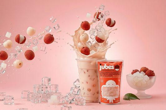 An action shot of lychee milk tea splashing out of a clear cup, surrounded by flying ice cubes, fresh lychees, and Jubes coconut gel, with a Jubes Lychee pouch and a bowl of nata de coco on a pink background.