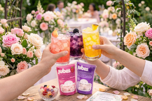 Three hands toast with colorful peach, blackberry, and pineapple mocktails filled with chewy Jubes coconut gel, set against a floral garden background with Jubes snack pouches on the table.