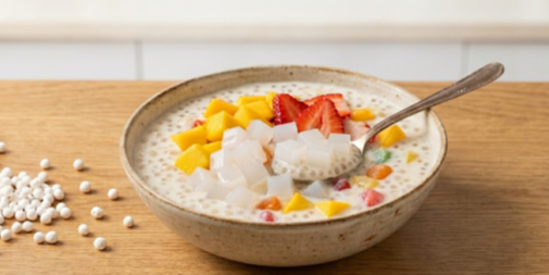 ceramic bowl filled with creamy sago pudding in coconut milk, topped with sliced mangoes, strawberries, and translucent white cubes of Jubes Nata de Coco. A silver spoon rests in the bowl, lifting a portion of the pudding and jellies.