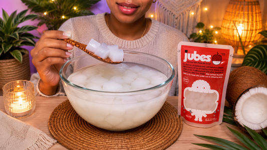 A woman lifts a spoonful of translucent coconut gel cubes from a large glass bowl, sitting next to a red pack of Jubes Lychee Flavor Nata De Coco and fresh coconuts on a warmly lit table.