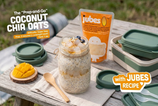 Mason jar of coconut chia oats topped with golden nata de coco cubes, blueberries, and coconut flakes, beside a mango half and an orange Jubes pouch.