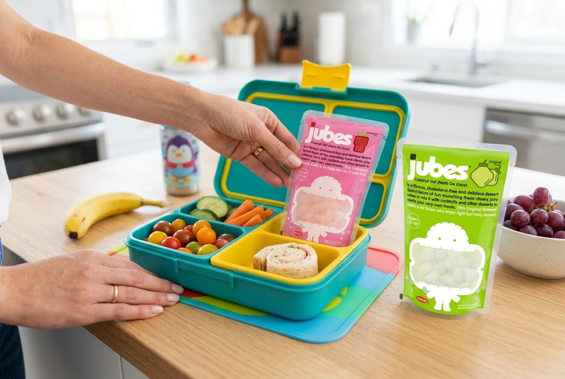 A hand places a pink Jubes Strawberry pouch into a kids' lunchbox with veggies and a sandwich, beside a green Jubes Original pouch and grapes.