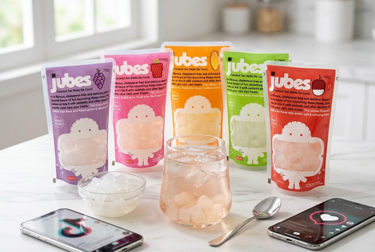 Five Jubes Nata De Coco pouches in Grape, Strawberry, Mango, Original, and Lychee beside a glass of iced coconut jelly drink and phones showing TikTok.
